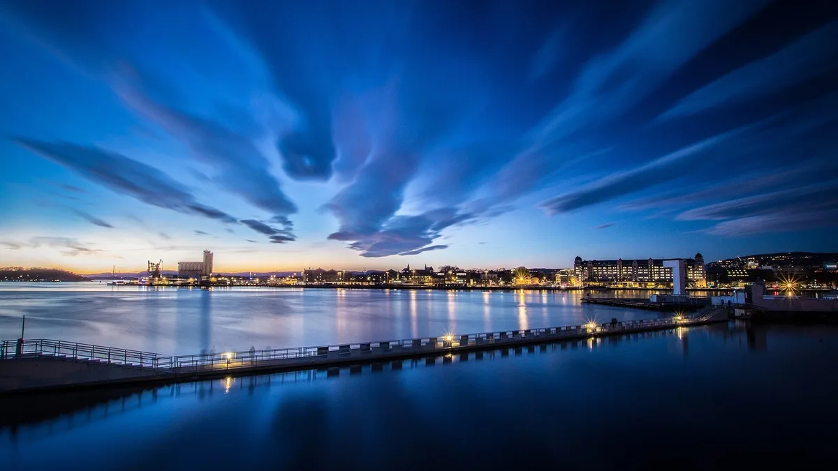 A long-exposure photograph of a city waterfront at dusk, showing streaked blue clouds, calm water reflecting city lights…