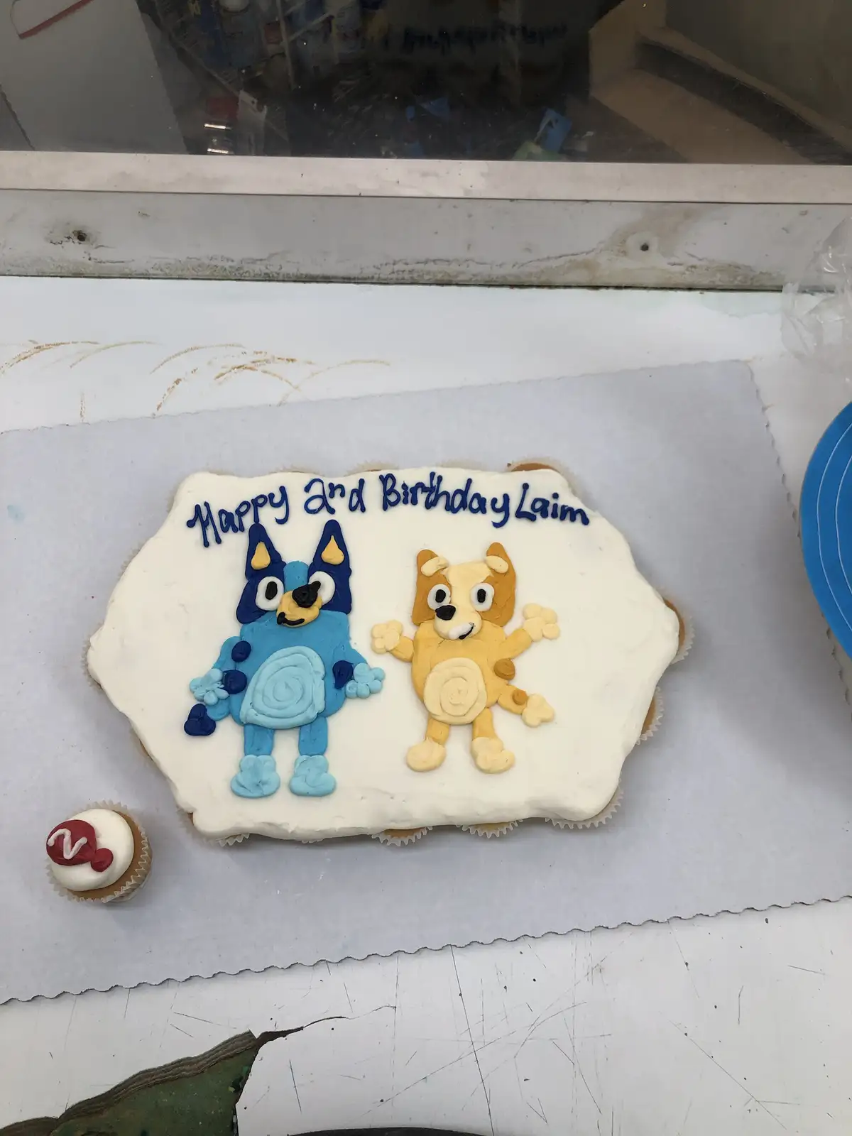 A rectangular cake with white frosting features two 3D fondant figures of the cartoon characters Bluey and Bingo from the…