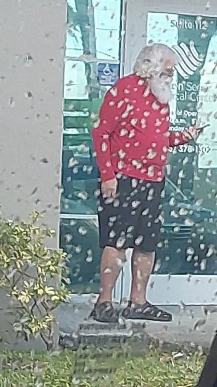An older man with a white beard and glasses stands outside a building, wearing a red long-sleeved shirt and black shorts…