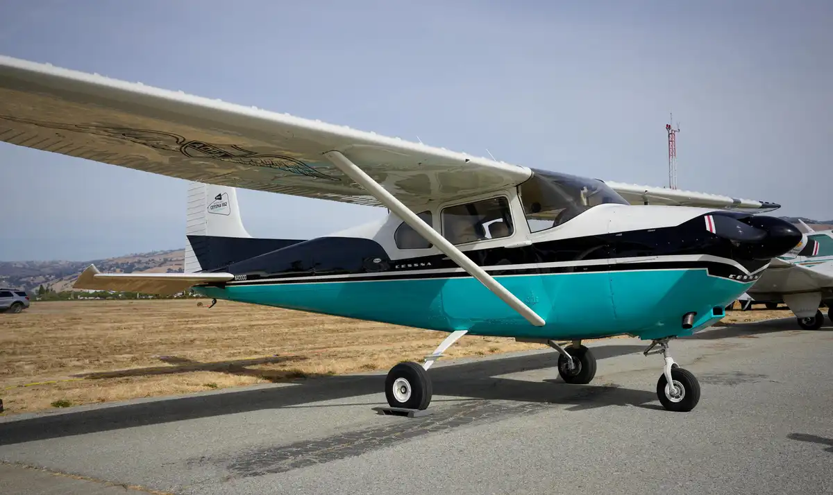 A Cessna 182 light aircraft with a white, black, and teal paint scheme is parked on an asphalt tarmac at an airfield, with…