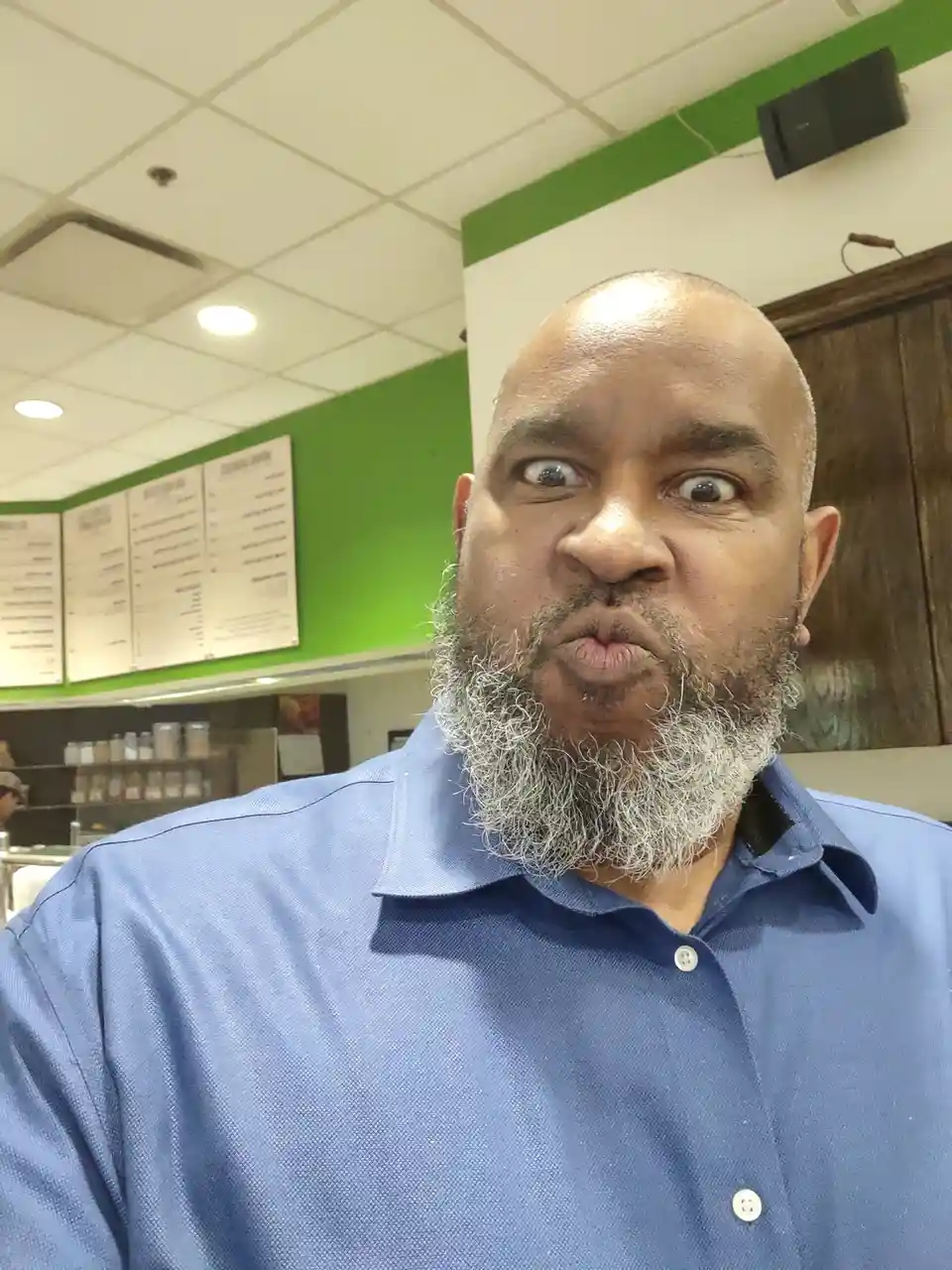 A man with a gray beard and blue shirt makes a comically disgusted face with wide eyes and puckered lips inside a coffee…