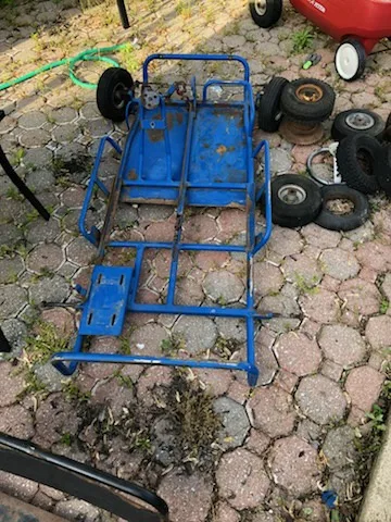 A blue, rusty, disassembled go-kart frame lies on a paved patio, surrounded by scattered tires and wheels. The overlaid text…