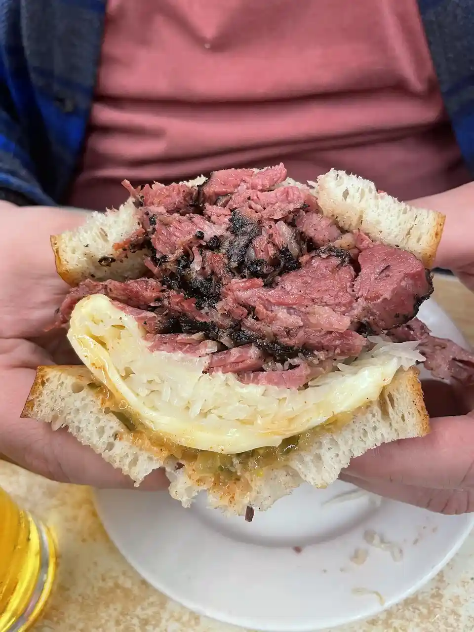 A person holds a generously filled sandwich with shredded pink meat, sauerkraut, and melted cheese between two slices of…