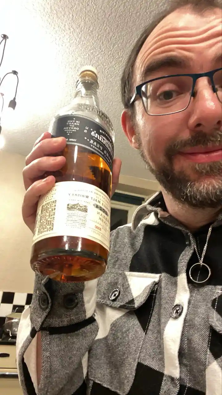 A man with glasses and a beard holds up a bottle of whiskey, looking at the camera with a slight smirk. The bottle has a…