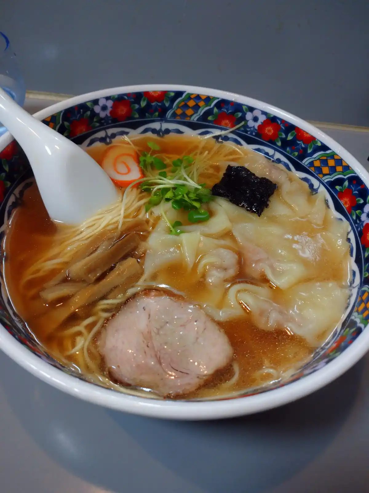 A close-up photo of a steaming bowl of ramen featuring a large slice of chashu pork, wontons, bamboo shoots, green onions…
