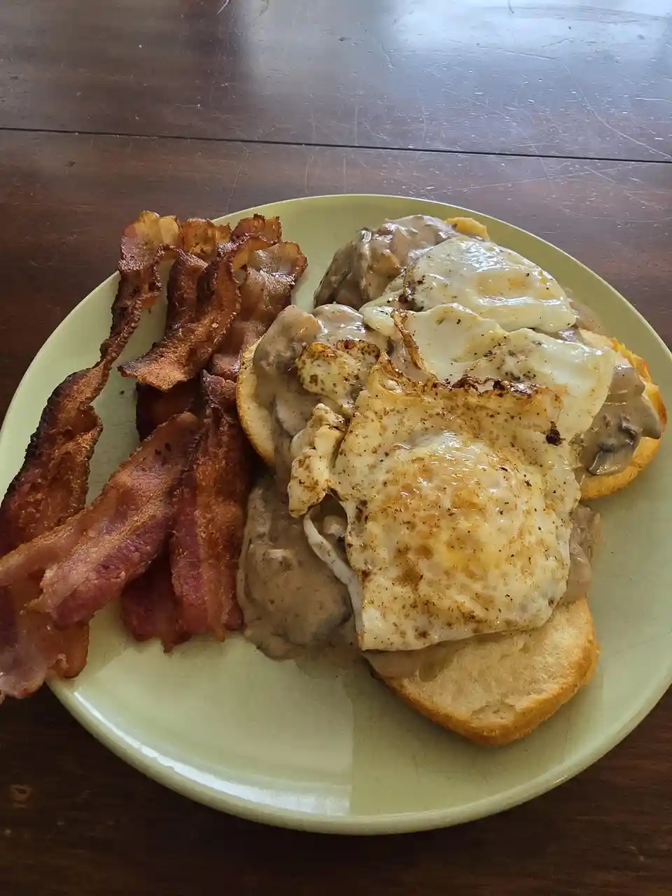 A plate of breakfast food featuring crispy bacon and a sandwich with fried eggs, mushroom gravy, and cheese on a bun, served…