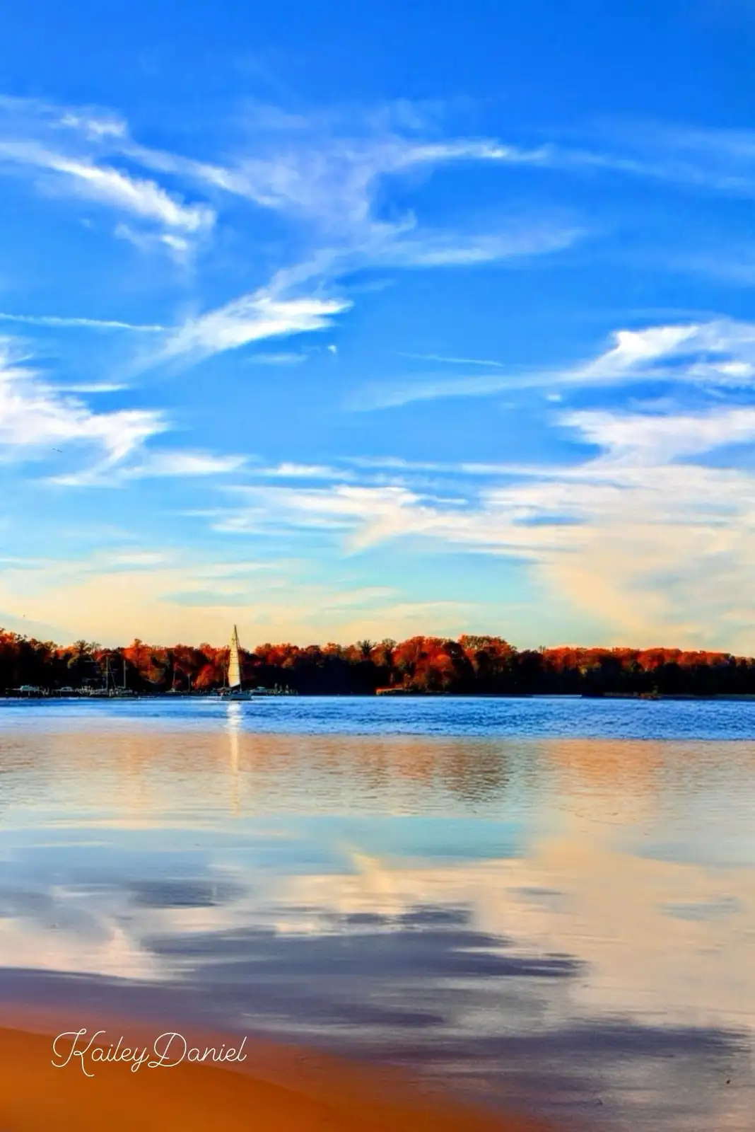 A serene landscape photo shows a sailboat on calm water with autumn-colored trees on the horizon under a bright blue sky…