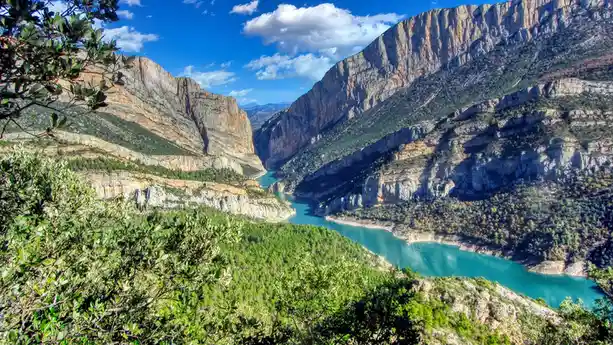 A scenic view of a turquoise river winding through a deep canyon with steep, rocky cliffs and green vegetation under a blue…
