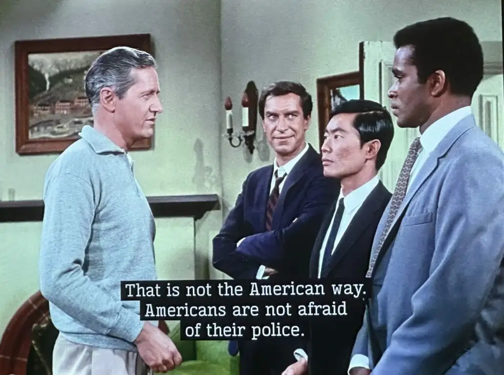 A still from a 1960s TV show shows four men in a room. One man in a grey sweater faces three other men in suits. Overlaid…