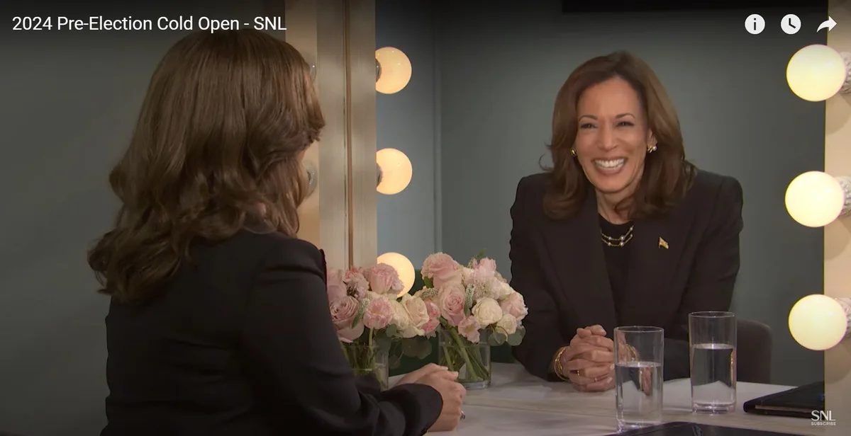 A woman in a black suit sits at a table smiling broadly while facing another woman whose back is to the camera; the scene…