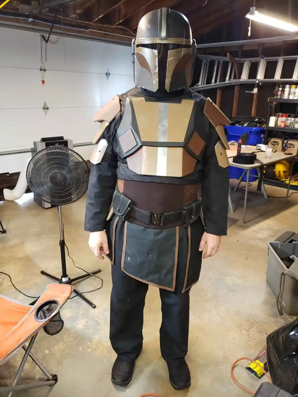 A person stands in a garage wearing a full Mandalorian armor costume, including helmet, chest plate, shoulder pads, belt…