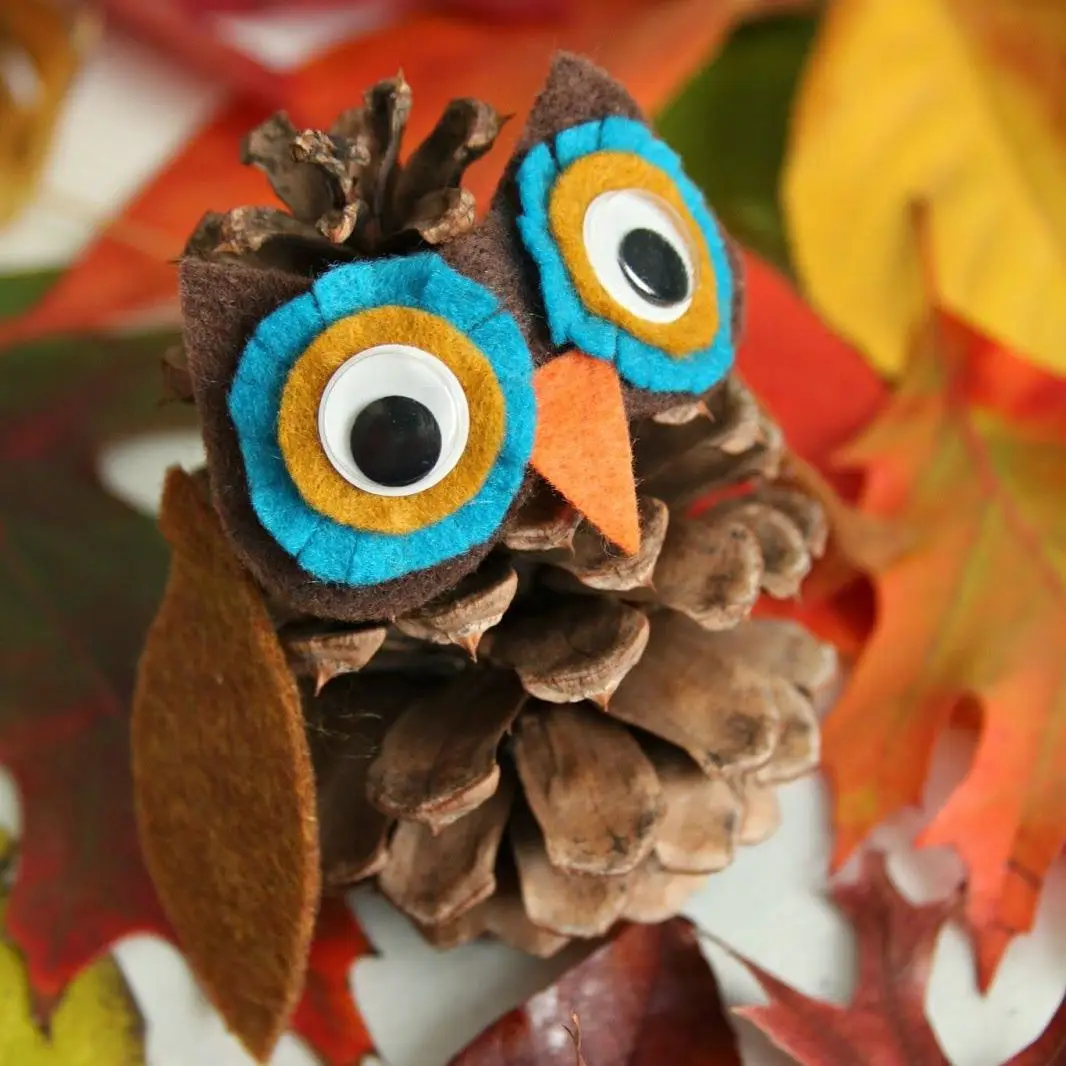 A close-up of a pinecone decorated with felt to resemble an owl, featuring large googly eyes with blue, yellow, and white…