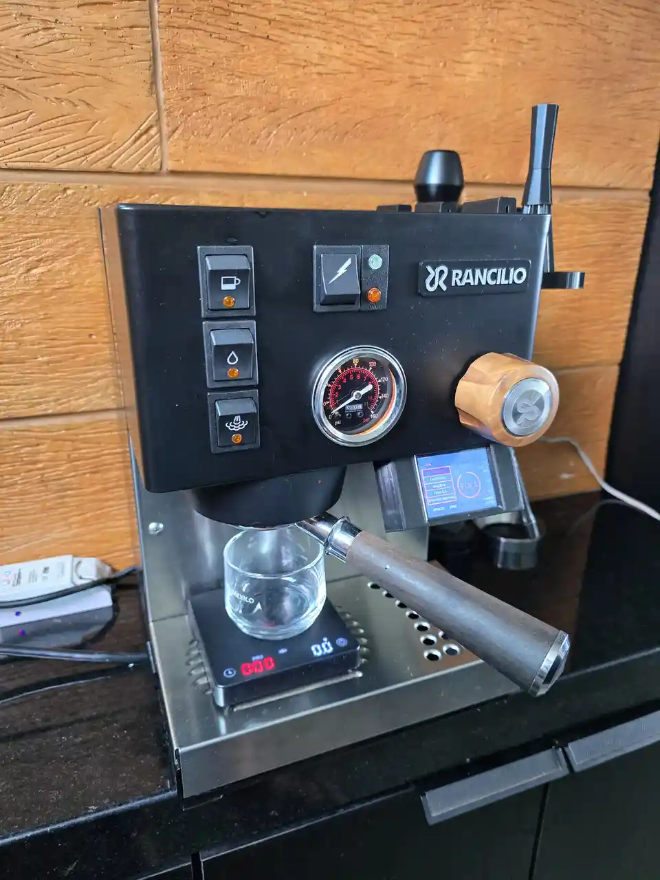A black Rancilio espresso machine sits on a dark countertop against a wood-paneled wall, with a glass cup on a digital scale…