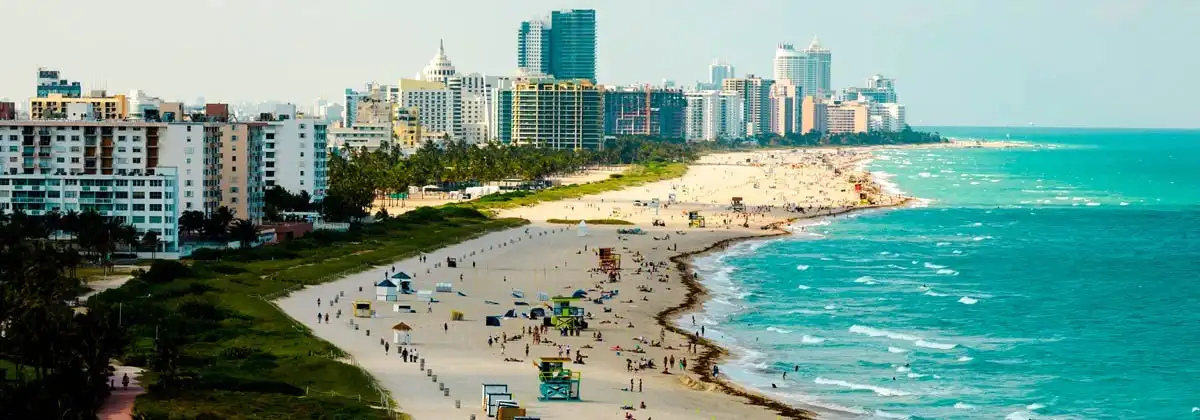 A wide-angle photo of a crowded beach with turquoise ocean waves and a dense city skyline of high-rise buildings in the…