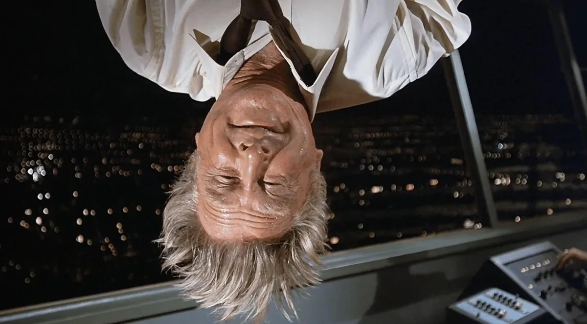 A man with gray hair and a white shirt is shown upside down, his face sweaty and eyes closed, seemingly in distress. Behind…