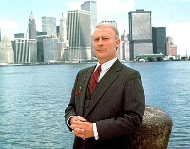 a man in a dark pinstripe suit and red tie stands with clasped hands against a waterfront backdrop featuring the New York…