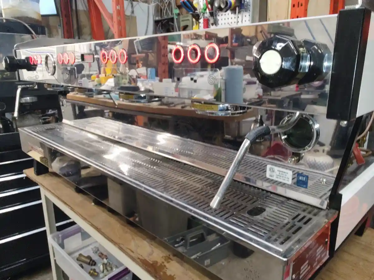 A large, shiny commercial espresso machine with red indicator lights is placed on a wooden workbench in a cluttered…