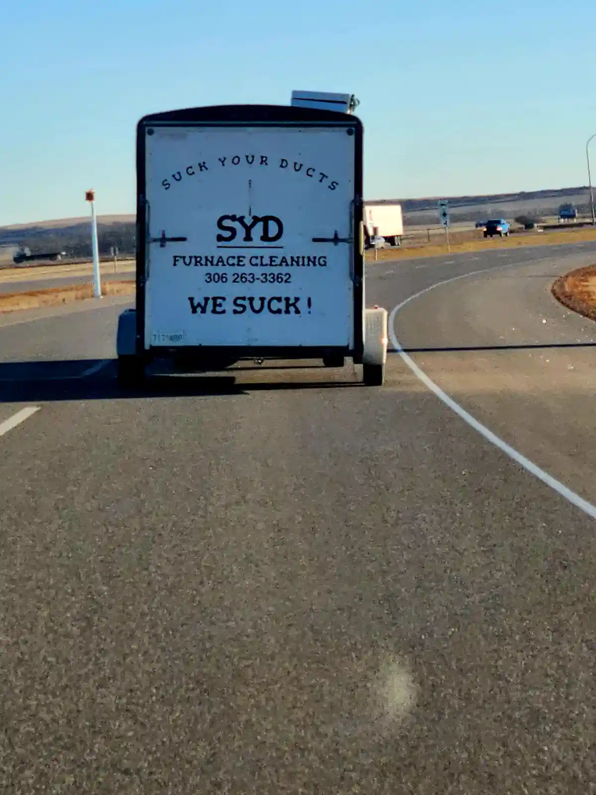 A white utility trailer on a highway has black text advertising 'SYD FURNACE CLEANING' with the phone number 306 263-3362…
