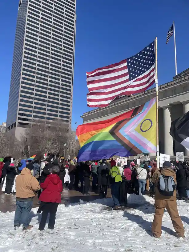 A crowd of people stands in snow-covered ground during a protest, with a large American flag and a rainbow flag (with a…