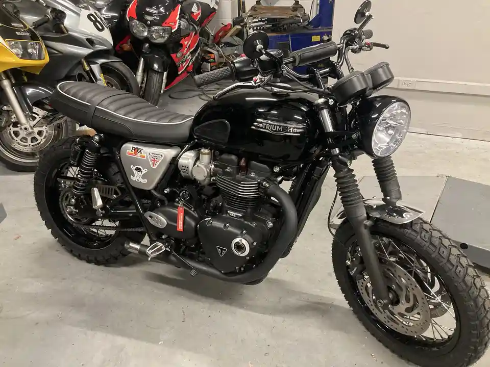 A black Triumph motorcycle with a classic cafe racer design, featuring a round headlight, black leather seat with white…