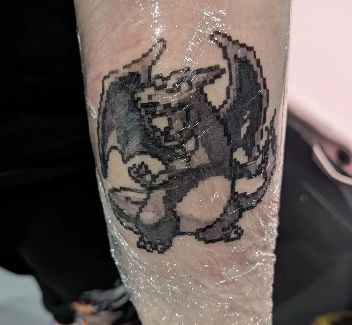 A freshly inked black-and-white pixel art tattoo of a Pokémon, resembling a Bulbasaur, is visible on a person's leg, covered…