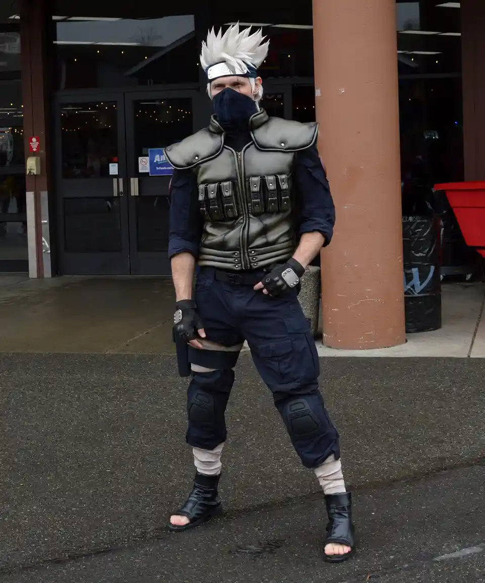 A man in a detailed cosplay of the anime character Kakashi Hatake stands outdoors, leaning against a pillar. He wears a dark…