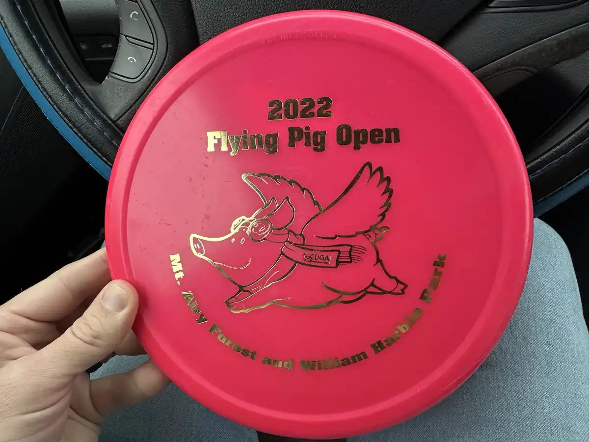 A person holds a bright pink disc golf disc inside a car, featuring a gold-foiled illustration of a flying pig wearing…
