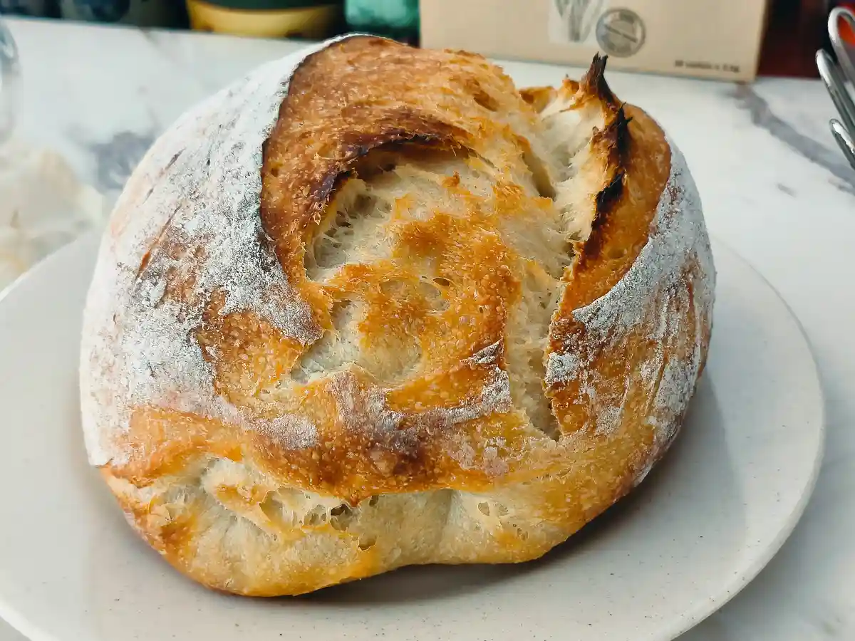 A round, rustic sourdough bread with a cracked, golden-brown crust sits on a white plate, dusted with flour. The top of the…