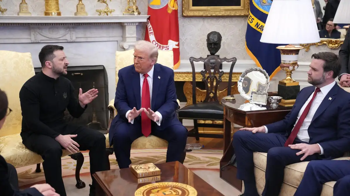 Three men sit in an ornate room with flags and a fireplace; the man in the center gestures while speaking, flanked by two…