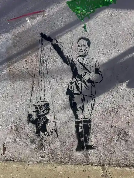 A black-and-white stencil graffiti on a textured wall shows a man in a military-style uniform holding puppet strings that…