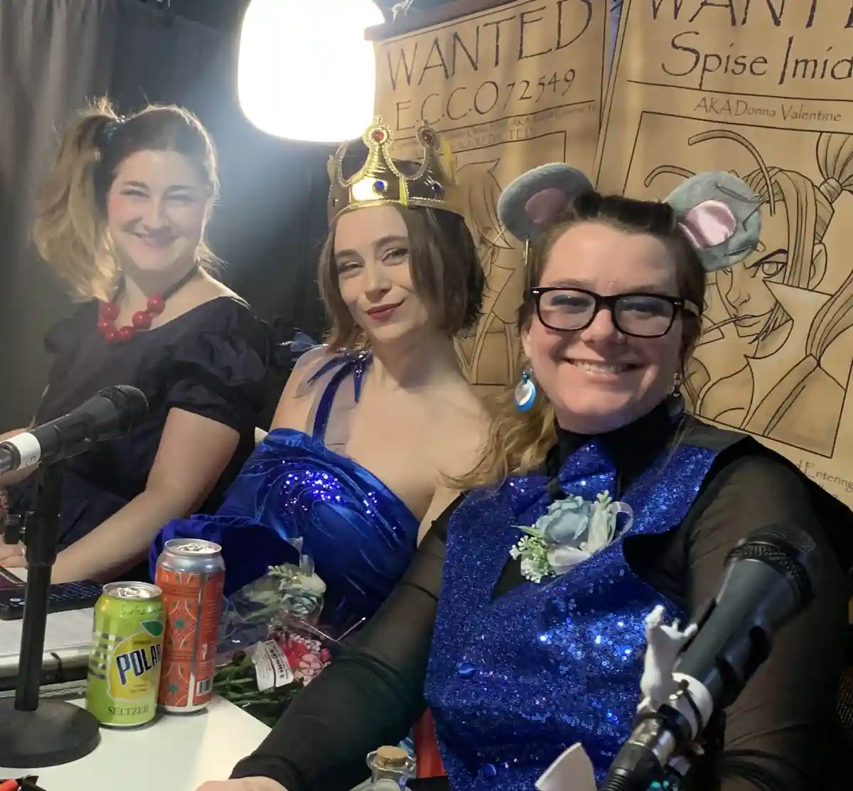 Three women in costumes sit at a table with microphones and drinks, with 'WANTED' posters behind them. The central woman…