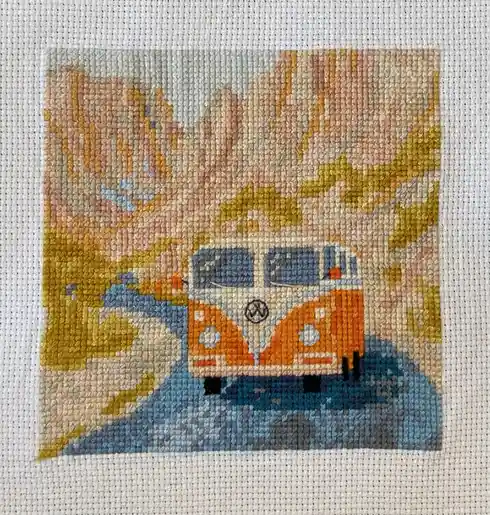 A close-up of a cross-stitch artwork depicting a vintage orange and white Volkswagen van parked on a blue path with stylized…