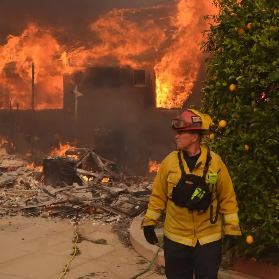 A firefighter in full gear stands calmly in front of a house engulfed in flames, with a citrus tree bearing fruit visible to…