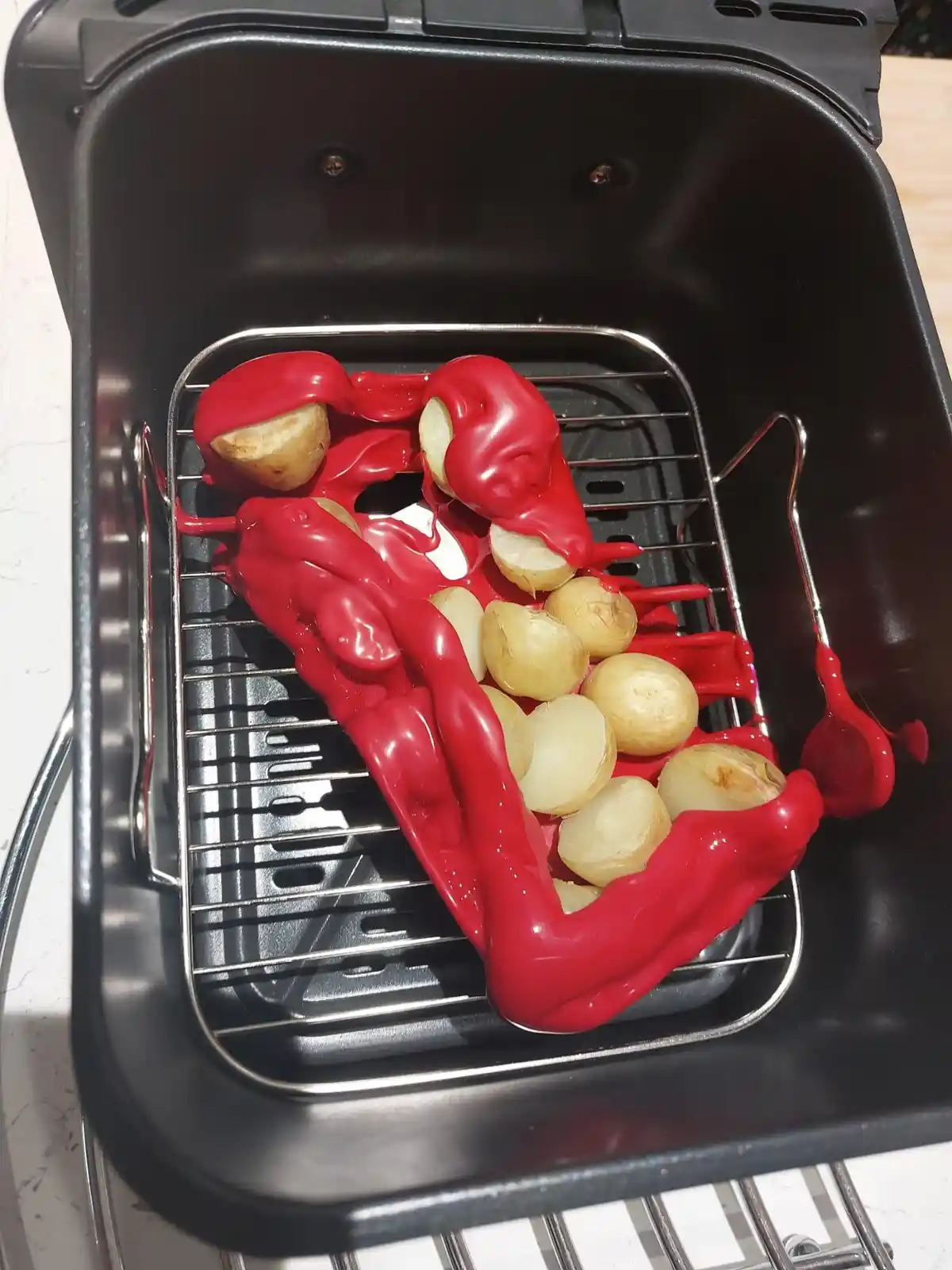 A black air fryer basket contains small boiled potatoes on a metal rack, partially covered by a thick, glossy red substance…