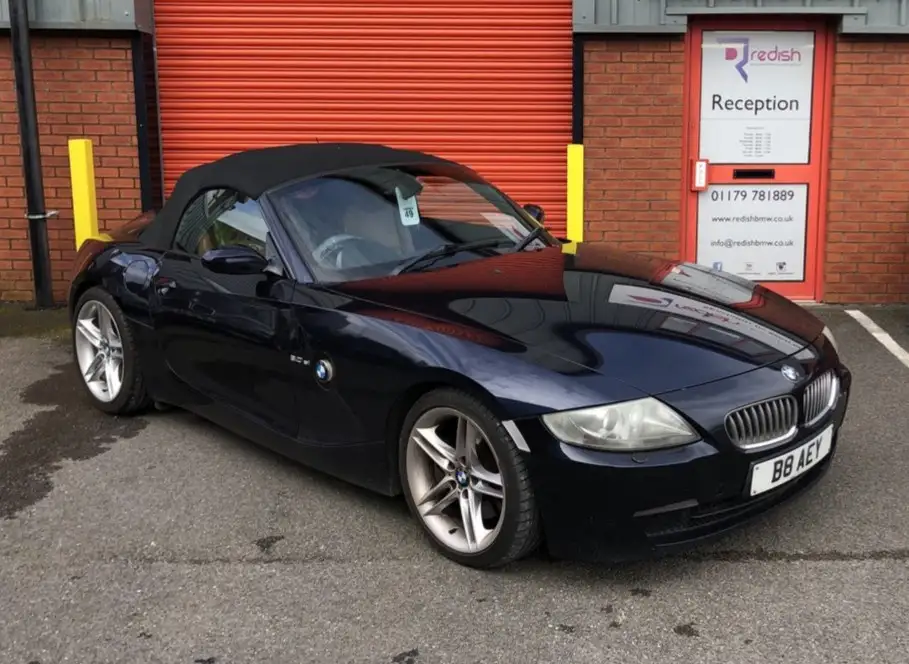 A dark blue BMW Z4 convertible is parked in front of a red brick building with a red roller door and a reception door. The…