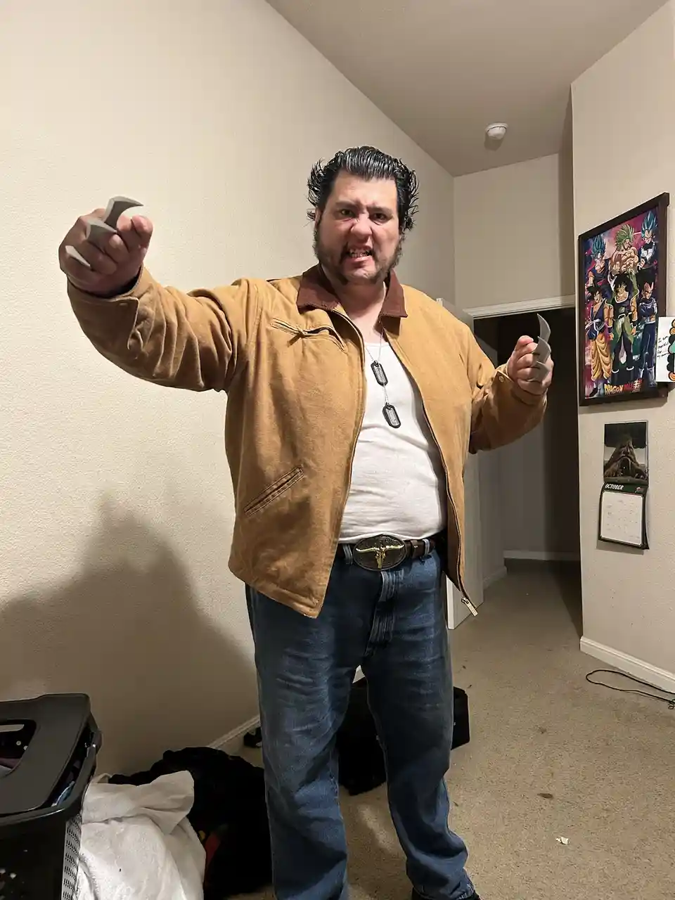 A man with a menacing expression, wearing a tan jacket and jeans, holds up two small, sharp, metallic claw-like objects in…