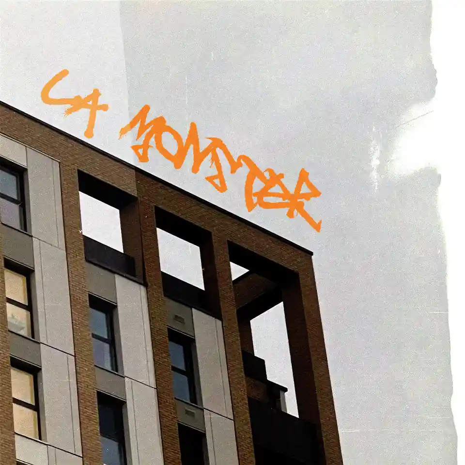 A low-angle shot of a modern apartment building with brown brick and gray panels, featuring orange spray-painted graffiti…