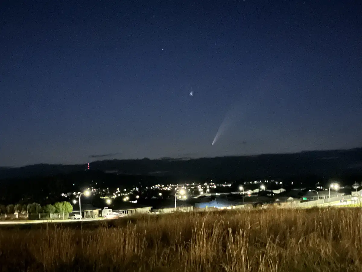 A night photograph shows a dark sky over a lit suburban area with hills in the background and tall dry grass in the…