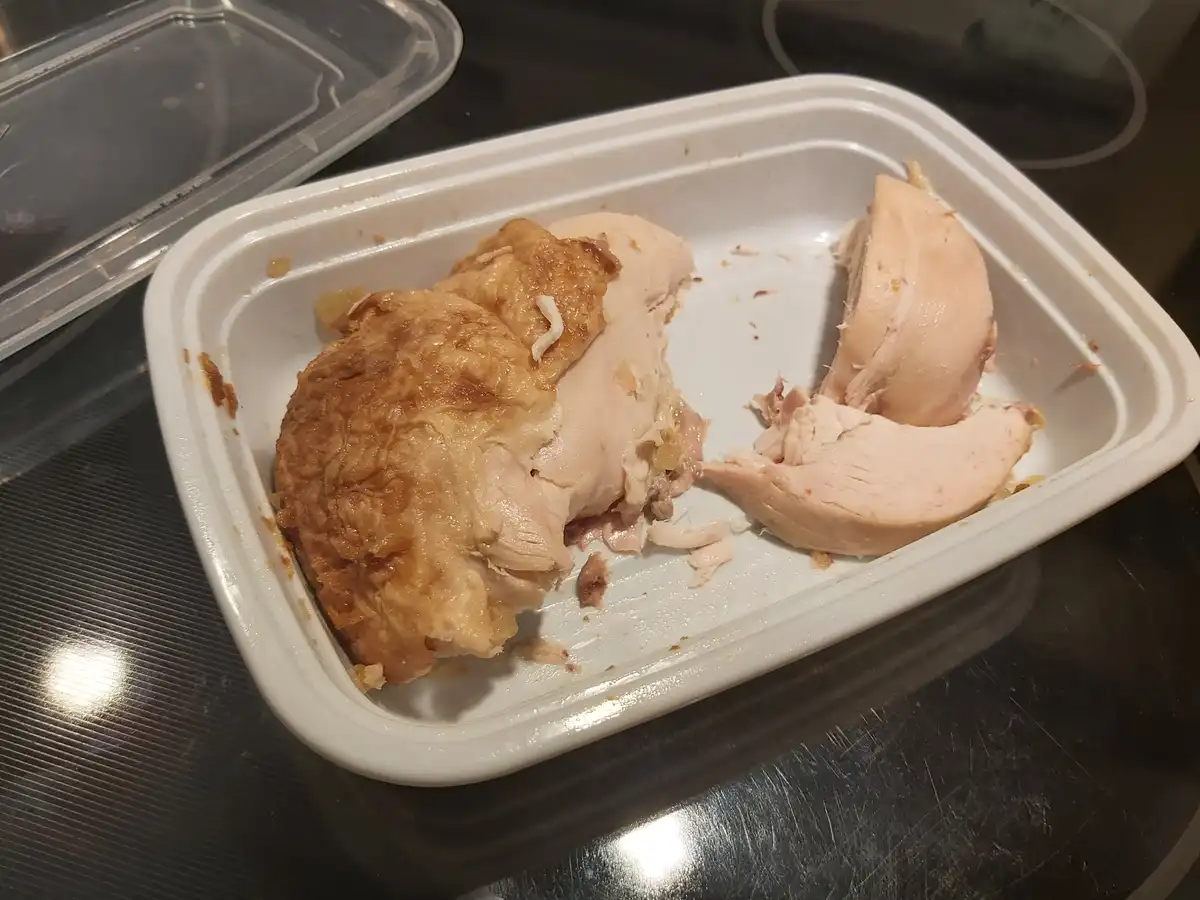 A white takeout container holds mostly eaten chicken, with one large, golden-brown fried piece and a few smaller, pale pink…