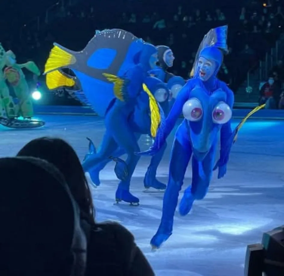 Ice skaters in blue Dory costumes from Finding Nemo perform on an ice rink, with one skater's costume featuring oversized…