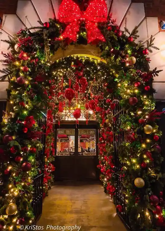 A grand, festive archway entrance to a building is heavily decorated with lush greenery, red and gold ornaments, twinkling…