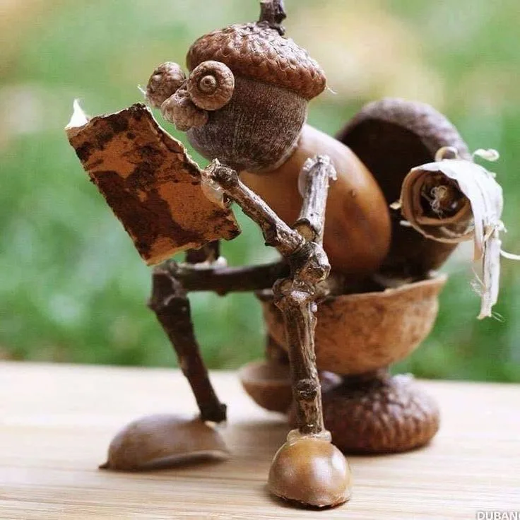 A whimsical sculpture made of acorns and twigs depicts a tiny creature sitting on a wooden surface, holding a small book…