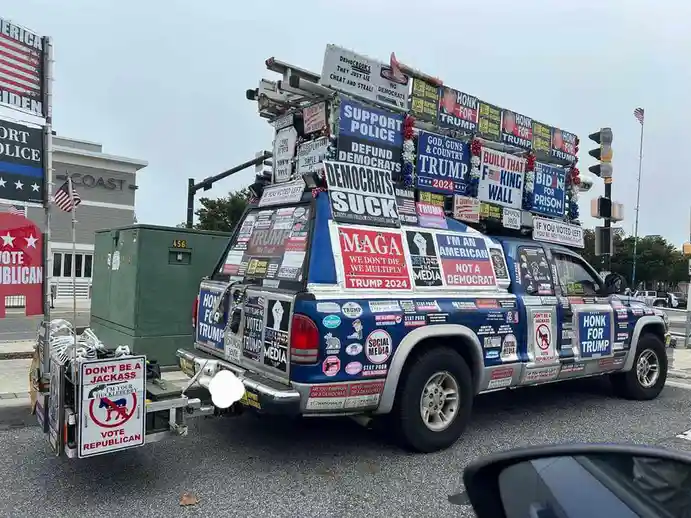 A pickup truck is covered in political signs and stickers, mostly supporting Donald Trump and criticizing Democrats, with a…
