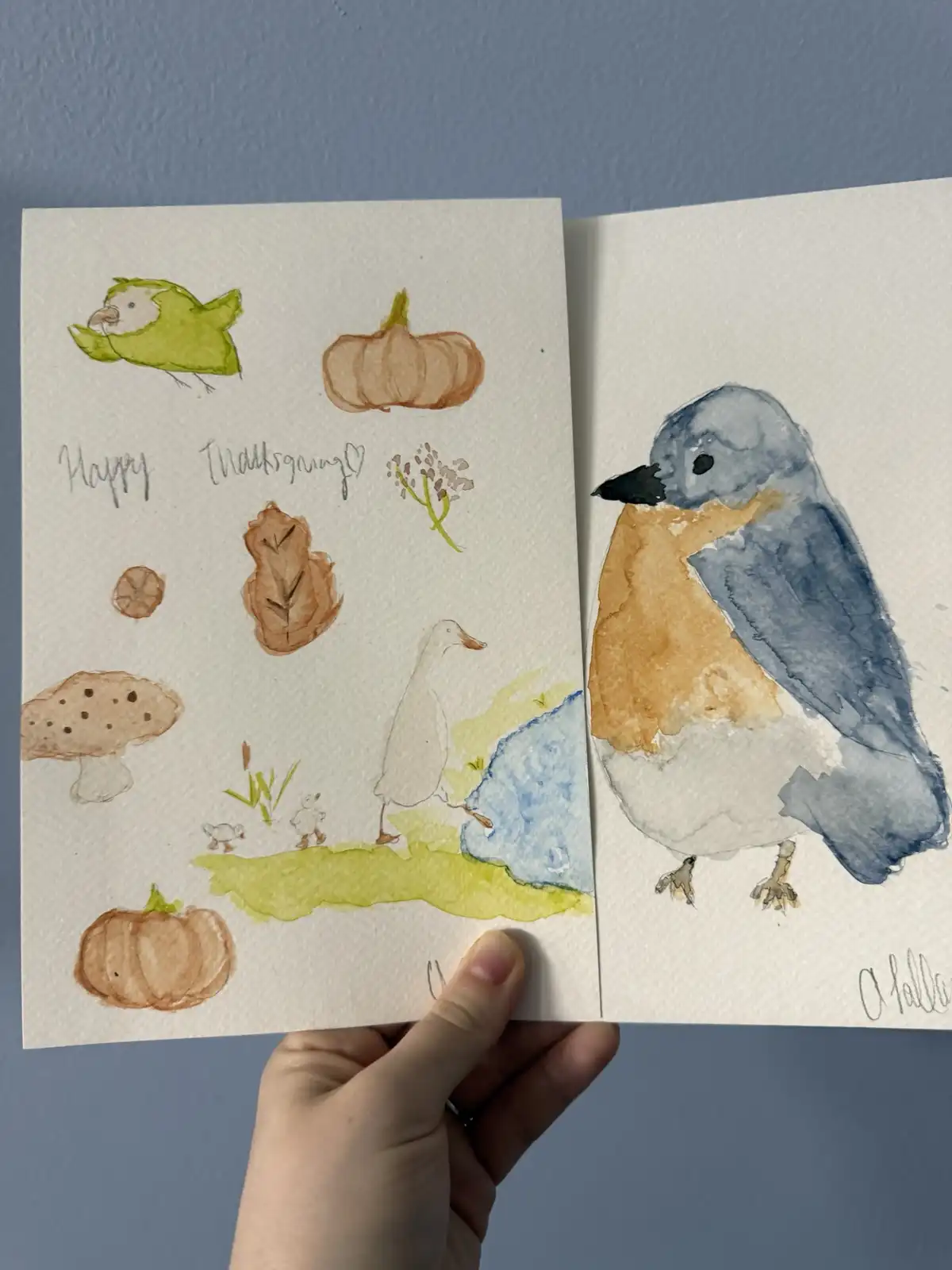 A hand holds two watercolor paintings side-by-side. The left page shows a festive Thanksgiving scene with pumpkins, a duck…
