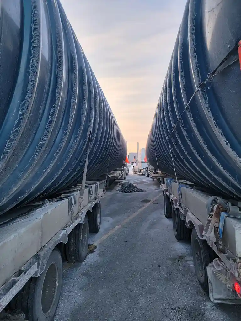 Two massive, dark blue corrugated pipes are loaded on flatbed trailers, facing each other to form a narrow corridor down a…