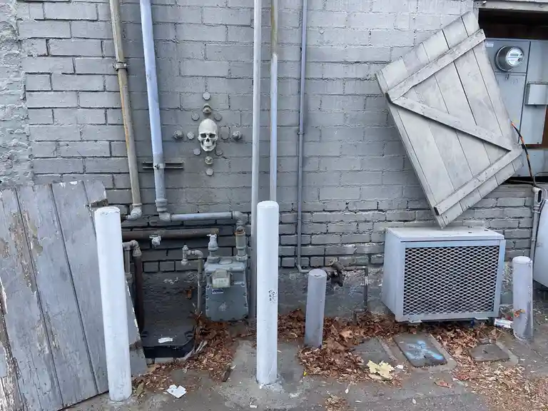 A gray brick wall features a white skull mounted among pipes and bolts, with weathered wooden planks leaning against it and…