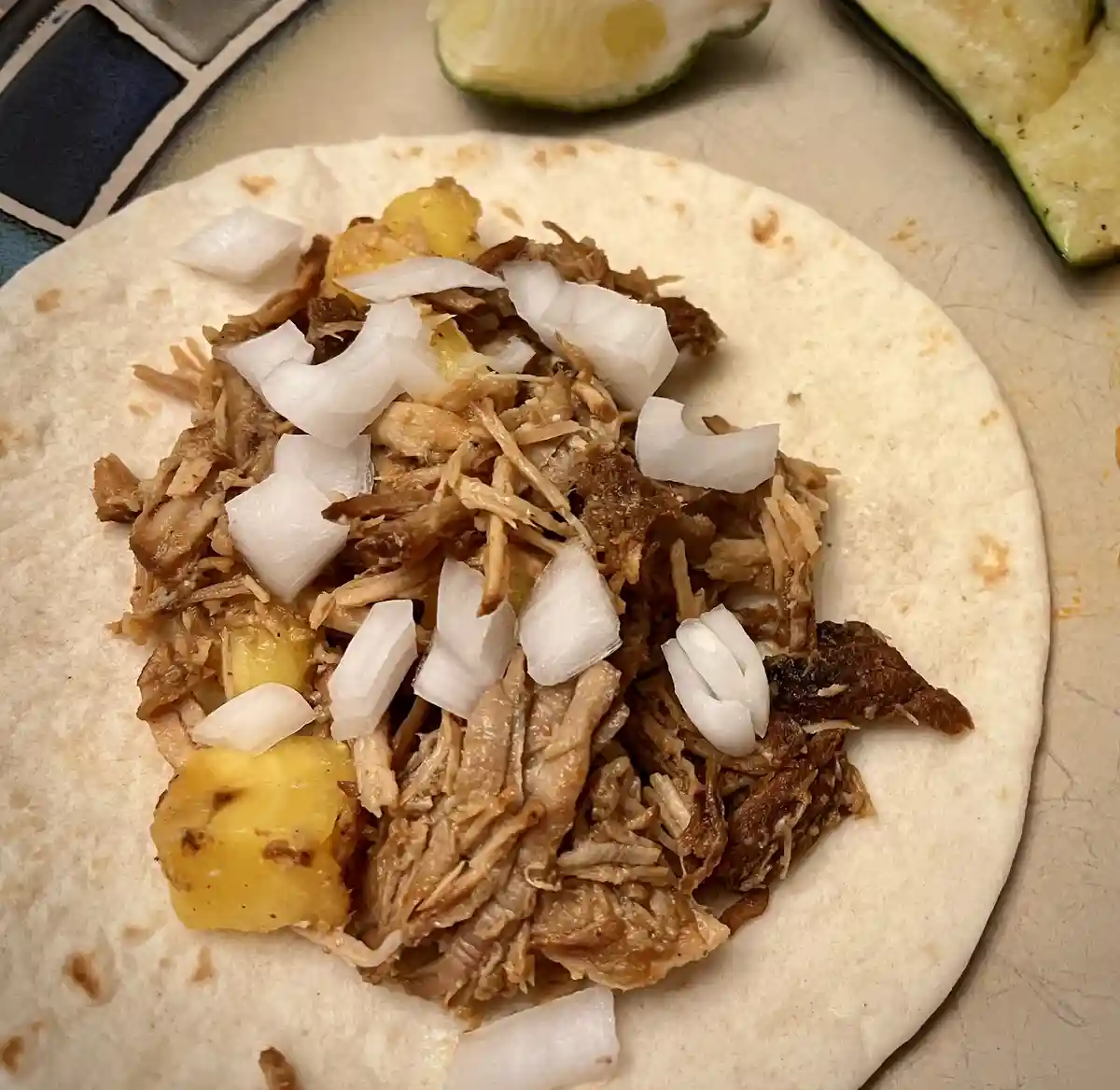 A close-up photo shows a soft tortilla topped with shredded meat, chopped white onions, and chunks of yellow pineapple. In…