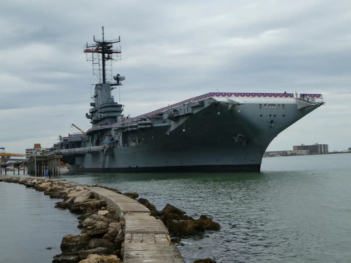 A large gray aircraft carrier is docked in a harbor under an overcast sky, with a concrete and rock breakwater in the…