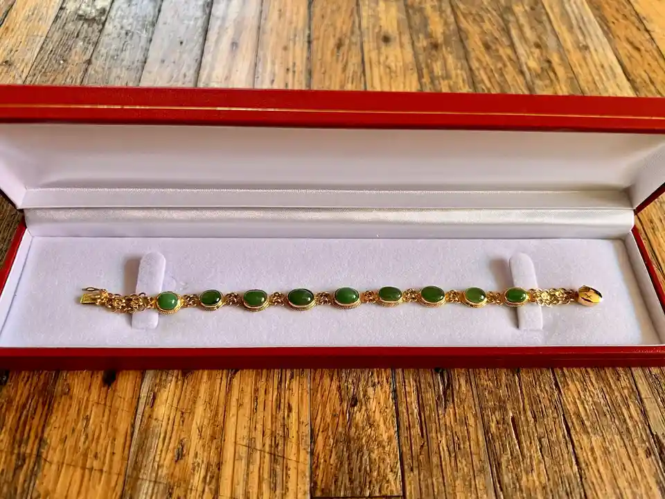 A gold bracelet with multiple green oval stones rests in a red jewelry box lined with white velvet, placed on a rustic…