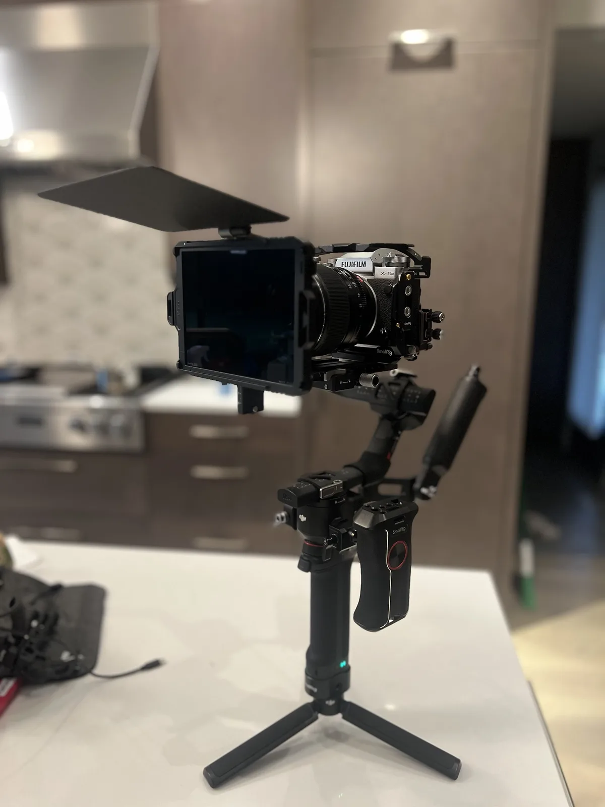 A Fujifilm X-T5 camera mounted on a DJI gimbal stabilizer with a large external monitor and a matte box, set up on a kitchen…